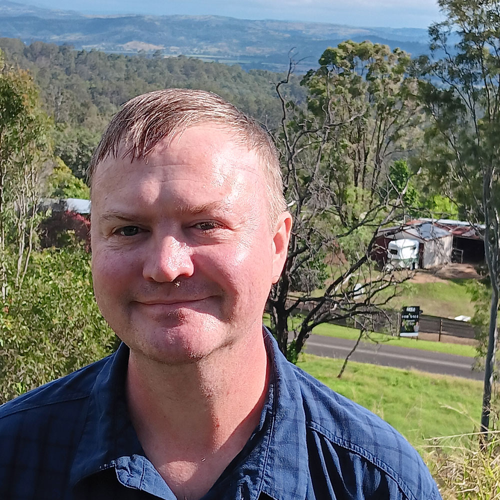 PhD candidate standing in a rural setting with paddocks, trees and hills stretching behind him