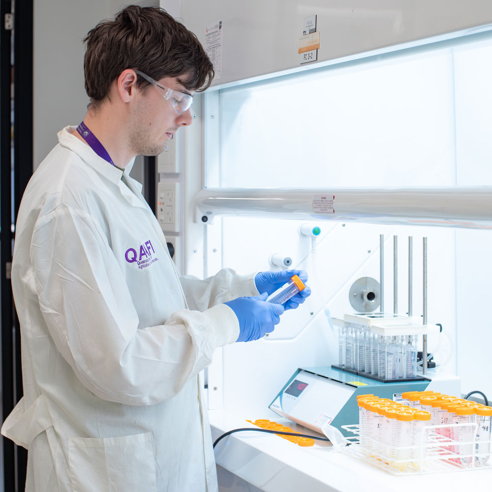 Man in laboratory setting wearing coat, gloves and glasses holding tube in a front of a machine