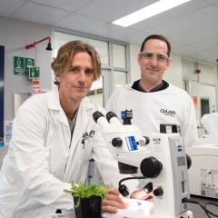 One scientists at a microscope and another standing behind him 